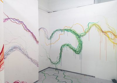 Photograph of immersive abstract painting in studio space. Three walls are visible, two to the left and one to the right making a corridor. The paint on the left is mainly bright blue and grey long lines, moving across the white walls. On the right wall is a large watercolour blue section, trailing into a warmer yellow and red, followed by a deeper green. The colour is trickling onto the floor, making lines moving from one wall to the next.